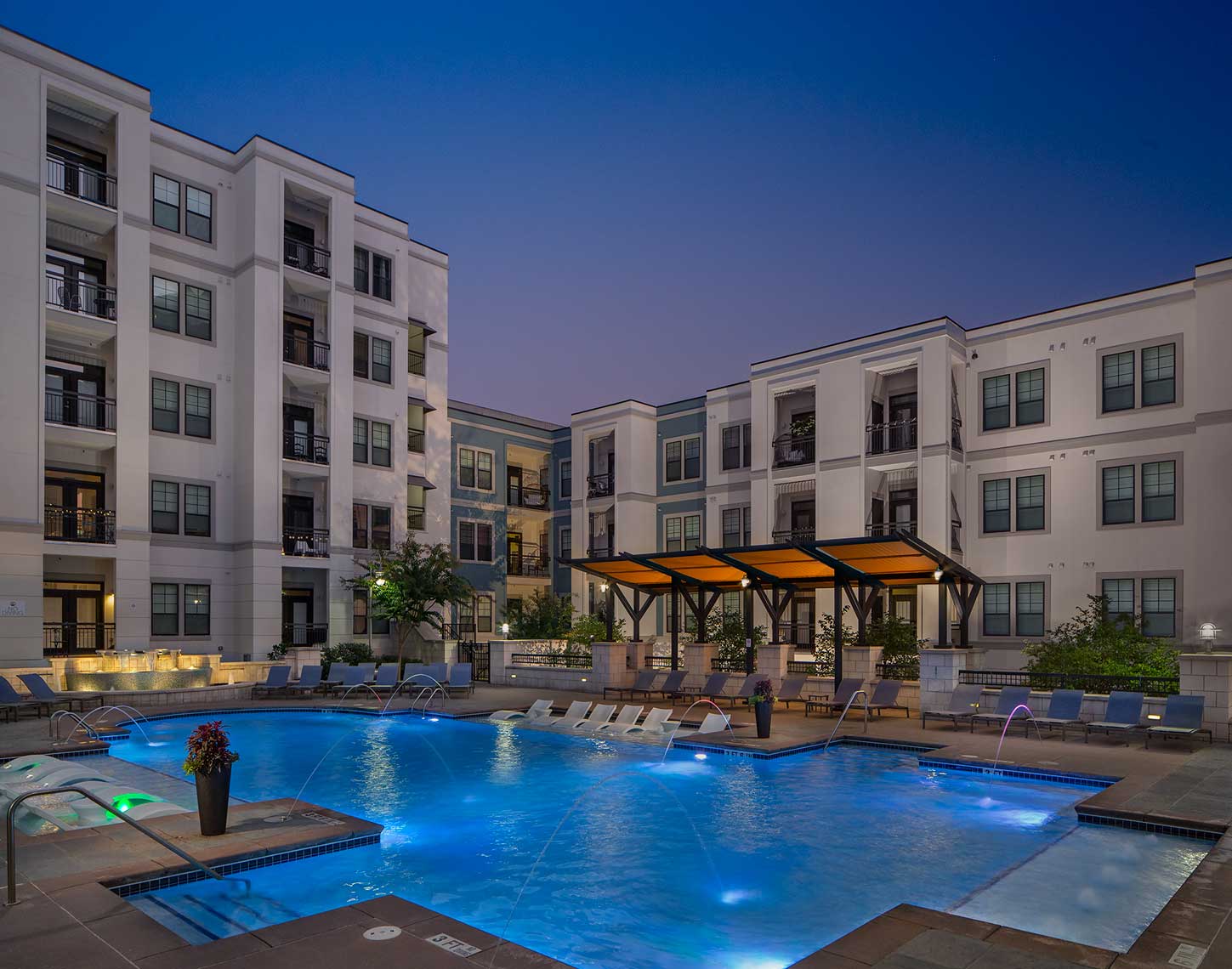 Dusk shot of pool with in-water lounge seats, additional lounge chairs, lighted fountain and covered area