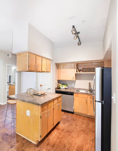 Modern open-style kitchen with track lighting, light brown cabinets, granite countertops with double sink, upgraded appliances and kitchen island on wood floors at MAA Uptown Village luxury apartments in Dallas, TX